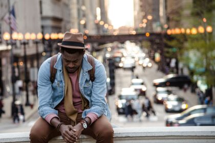 A man in a leather jacket looking down while sitting on a ledge in a city